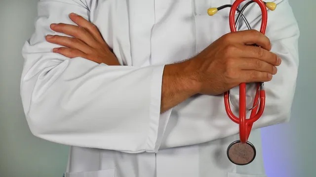 Profissional de saúde com jaleco branco segura um estetoscópio vermelho, representando a atuação médica